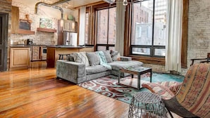 A spacious living area with a wooden floor, a sectional sofa, a coffee table, and a wicker chair. There is a kitchen area with cabinets and a refrigerator in the background.