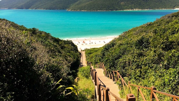Un escalier en bois mène à une plage bordée d’une eau turquoise, avec des collines verdoyantes en arrière-plan.