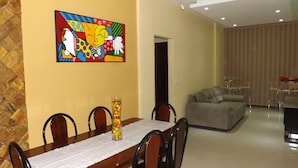 Un coin repas avec une table et des chaises en bois, une peinture abstraite aux couleurs vives accrochée au mur, et un vase en verre rempli de fruits posé sur la table.