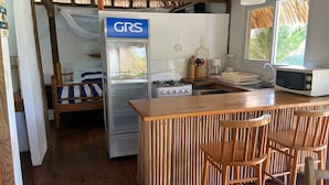 Uma cozinha compacta com fogão a gás, geladeira e micro-ondas. Possui um balcão de madeira com banquetas e uma janela que traz luz natural.