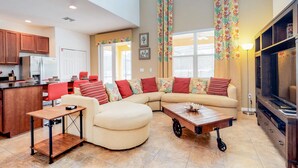 A modern living room with a sectional sofa, a coffee table, and a TV. There is a kitchen area with wooden cabinets and a refrigerator.