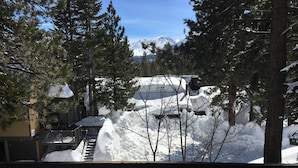 Schneebedeckte Landschaft mit einem Berg im Hintergrund, einer Holzdielen-Terrasse und einem Gebäude mit verschneitem Dach.