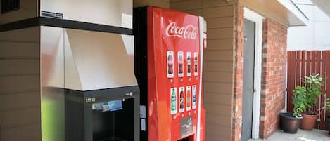 Una máquina expendedora de Coca-Cola y una zona de parrilla al aire libre con pared de ladrillo y plantas en macetas.
