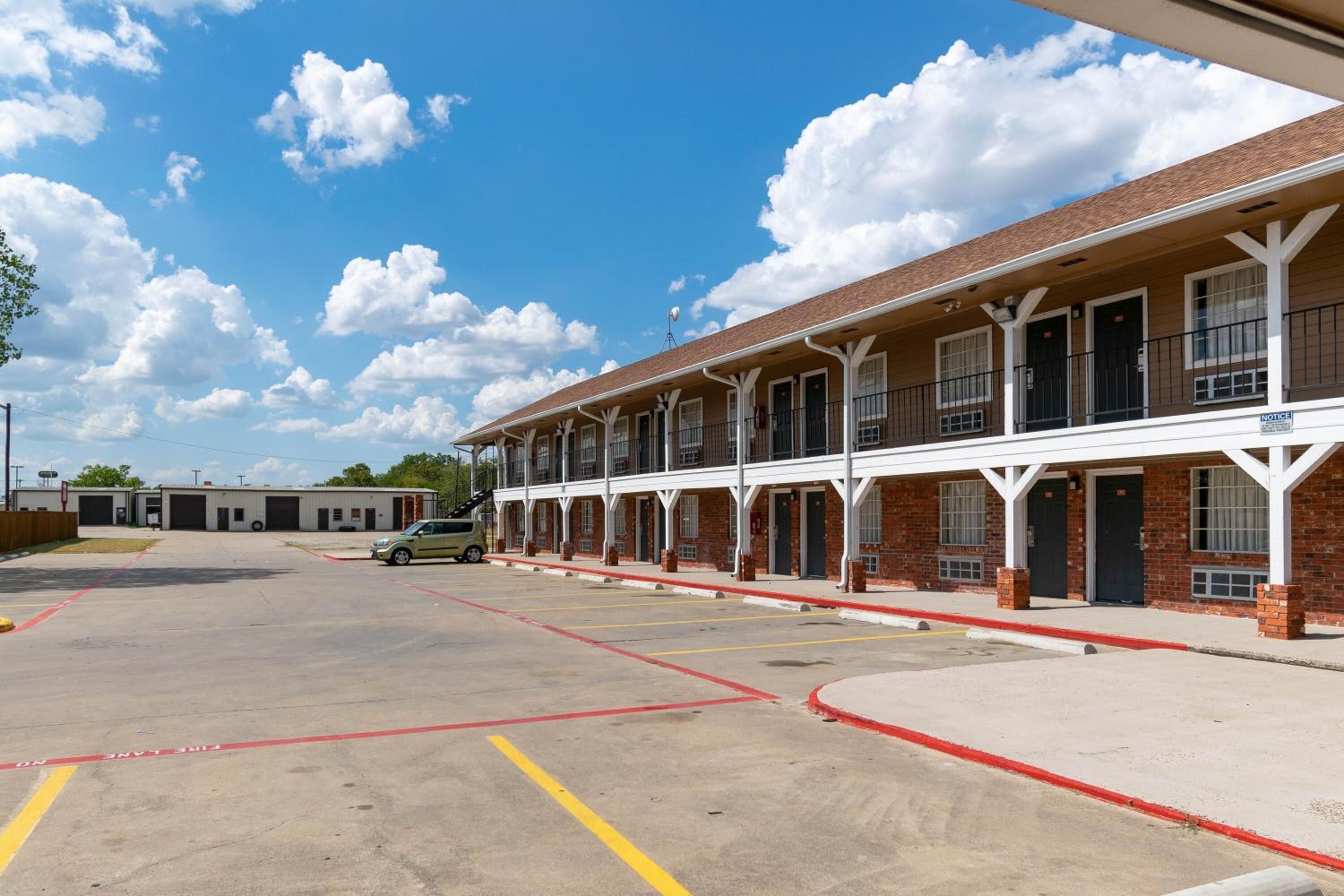 A motel with a row of rooms, a parking lot, and a building in the background.