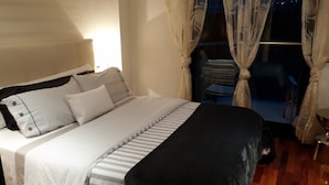 Un lit bien fait, avec une literie à rayures et un plaid noir. La pièce contient également une chaise et une petite table sur laquelle se trouve un bol.