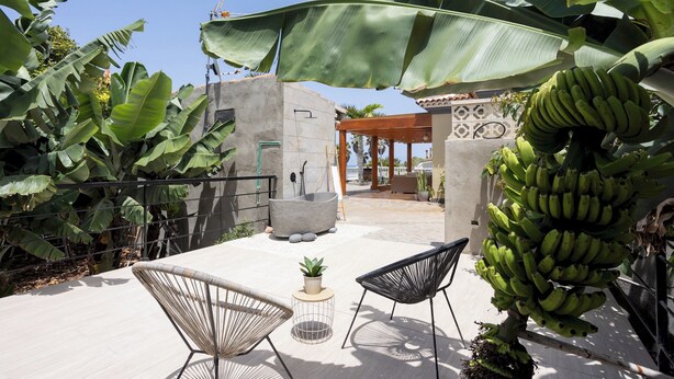 A patio area with two chairs, a small table with a plant, and a large banana tree with unripe bananas. - Home2Book Garachico Ocean View & Wifi (Las Cruces)