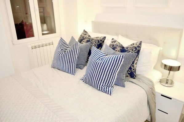 Una cama bien hecha con edredón blanco y varios almohadones, una mesita de noche con lámpara y una ventana con vistas al exterior.
