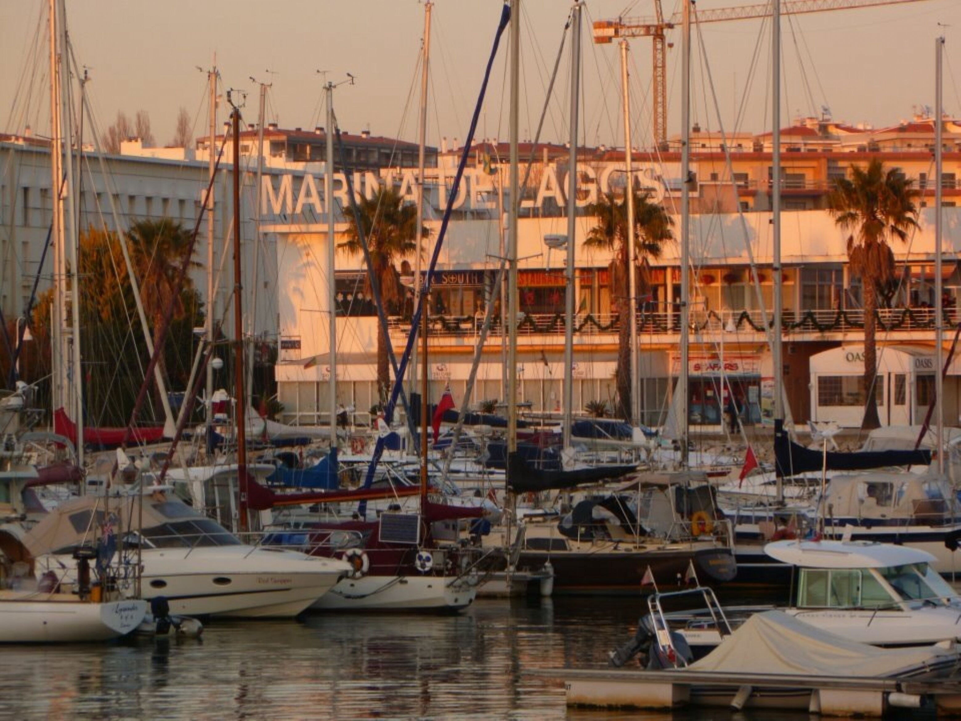 A marina with multiple boats docked, a building with 'MARINA DE LAGOS' signage, and palm trees in the background.