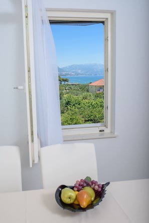 Una habitación con ventana que da a un paisaje costero, una mesa con frutero y una silla blanca.