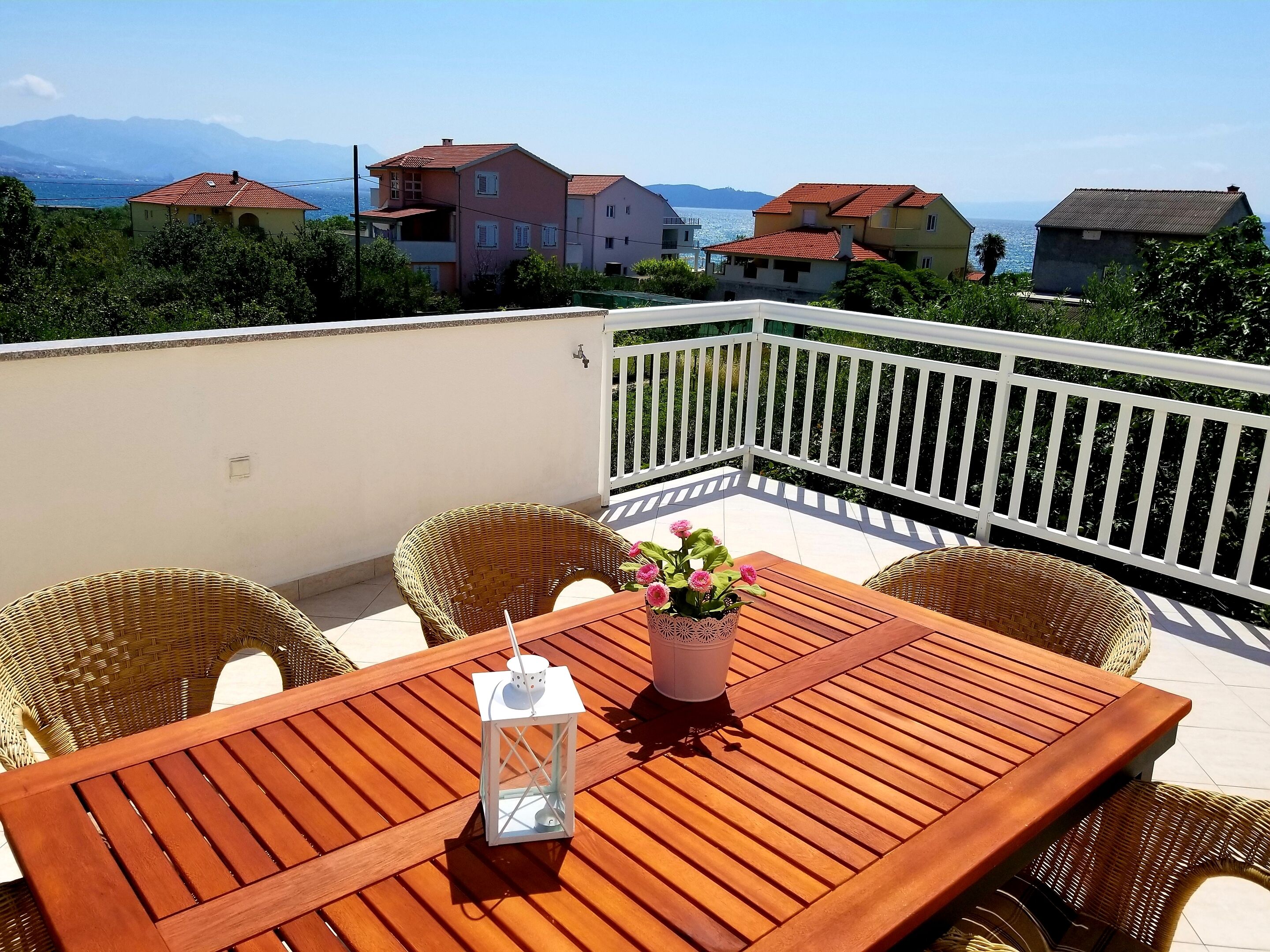 A wooden table with wicker chairs on a balcony with a white railing, overlooking a coastal area with houses and mountains in the background.