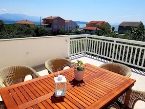 Una mesa de madera con sillas de mimbre en un balcón con barandilla blanca, con vistas a una zona costera que incluye casas y montañas al fondo.