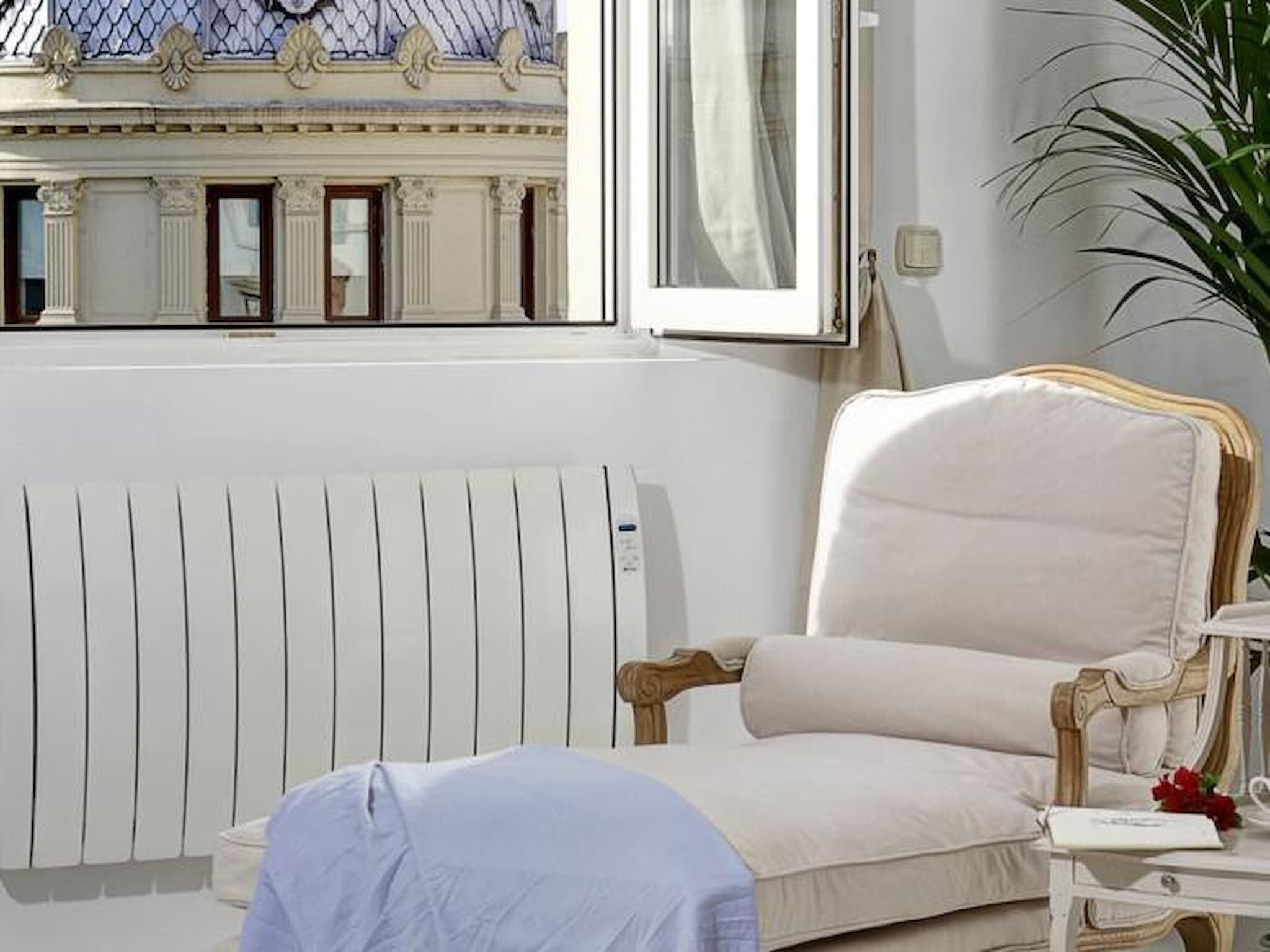 Una habitación con radiador, ventana con vista a un edificio, una silla y una mesita de noche con un libro y una flor.