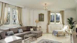 A modern living room with a sectional sofa, a coffee table, a chair, a lamp, and a view of the city through large windows. - ChezmoiHomes La Belle Epoque (Granada)