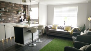 Een moderne keuken met een centraal eiland, barkrukken en een grijze bank met gele kussens in een woonruimte.