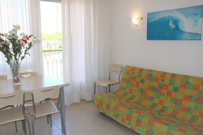 Una habitación con una mesa de comedor, sillas, un sofá, un cuadro de un surfista sobre una ola y una ventana con cortinas.
