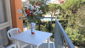 Un balcó amb una taula i caires blancs, un vas de flors i vista a una piscina i vegetació.