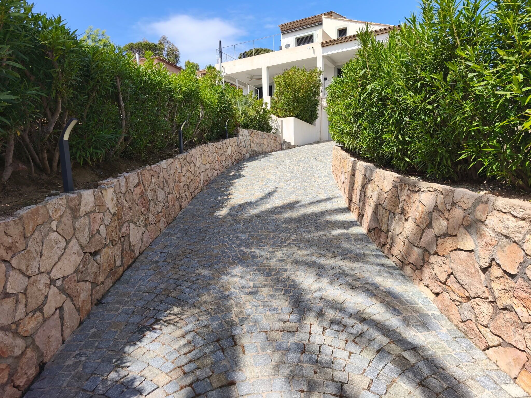 Un sentier pavé de pierres, bordé de murs en pierre et de verdure, mène à un bâtiment moderne doté d’une façade blanche et d’un toit brun.