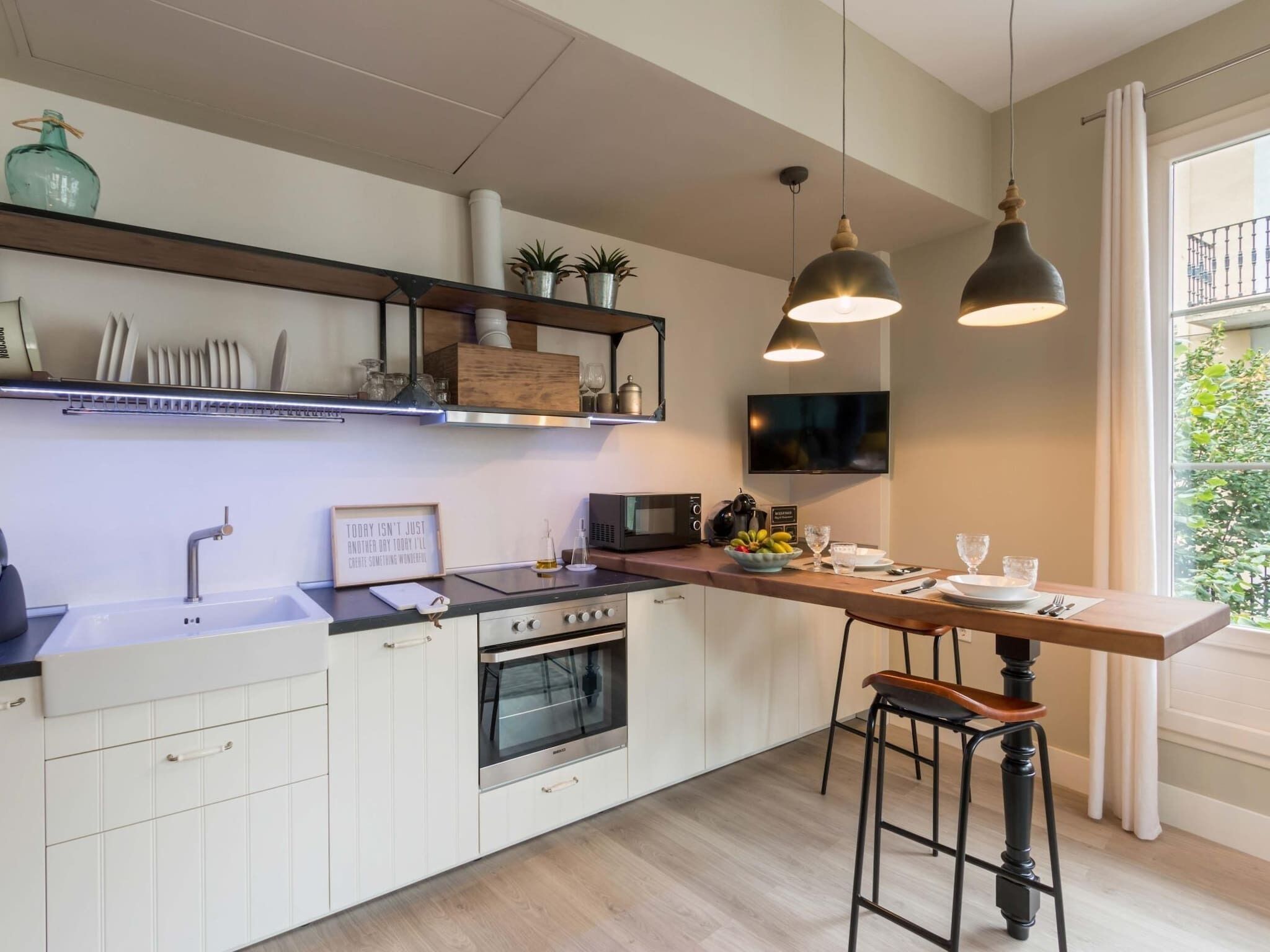 Una cocina moderna con armarios blancos, horno empotrado, microondas y cafetera. Hay dos taburetes de bar, una mesa de madera y un amplio ventanal con vistas a la vegetación.