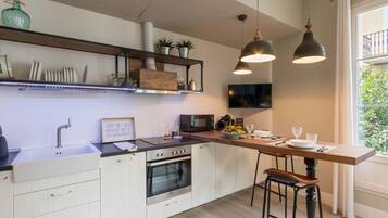 Una cocina moderna con armarios blancos, horno empotrado, microondas y cafetera. Hay dos taburetes de bar, una mesa de madera y un amplio ventanal con vistas a la vegetación.