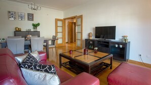 A modern living room with a sofa, coffee table, and TV. There is a dining area with chairs and a table. The room has a wooden floor and white walls. - CARCAVELOS BEACH 2. A wonderful 2 bedroom (Carcavelos)
