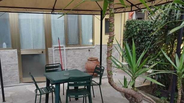 En utomhusplats med ett bord och stolar, en stor krukväxt och en pergola som ger skugga.