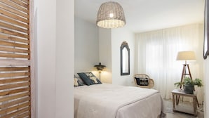 Un dormitorio con cama, lámpara de mesita de noche, lámpara colgante de mimbre, una silla, una mesita con una planta y un espejo en la pared.