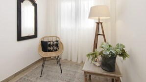 Una habitación con una silla de mimbre, una mesa con una lámpara y una planta en maceta, un espejo y una ventana con cortinas blancas.