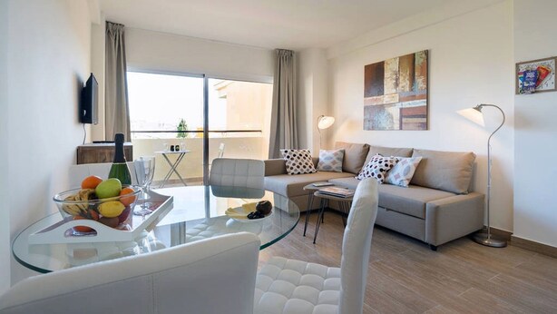 Un salon moderne avec une table à manger en verre, un canapé et une table basse. Il y a un balcon avec vue, un téléviseur fixé au mur et un tableau au-dessus du canapé.