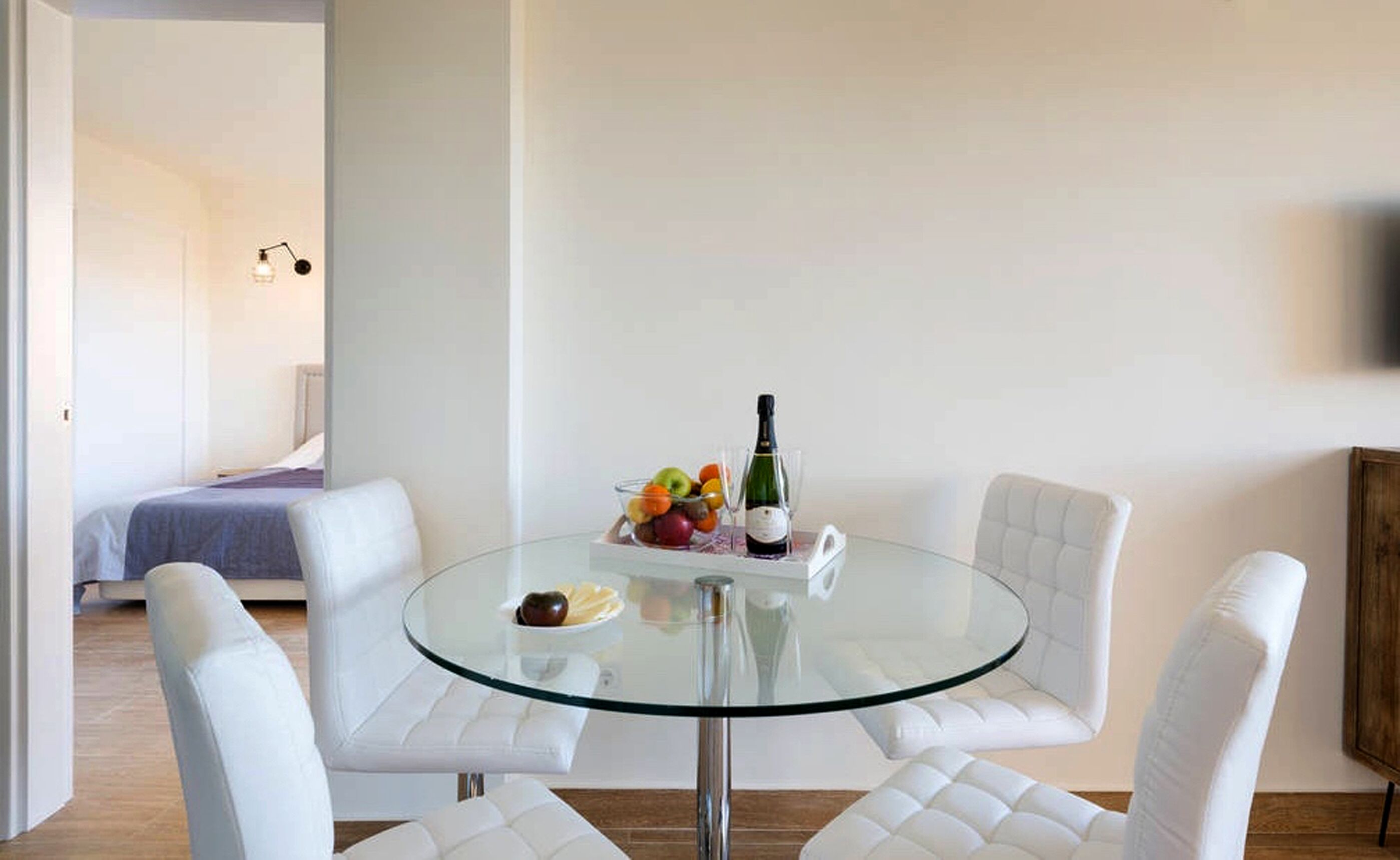 A modern room with a glass table, white leather chairs, a bed with a blue blanket, and a fruit bowl with a bottle on the table.