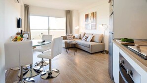Un salon moderne comprenant un canapé, une table à manger avec des chaises, un téléviseur à écran plat fixé au mur et un coin cuisine avec un réfrigérateur et une planche à découper.