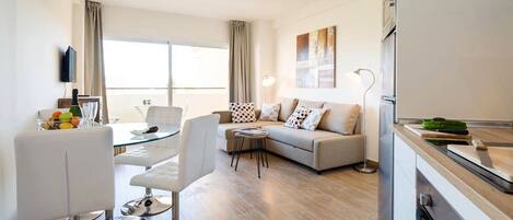 A modern living room with a sofa, a dining table with chairs, a flat-screen TV mounted on the wall, and a kitchen area with a refrigerator and a cutting board.