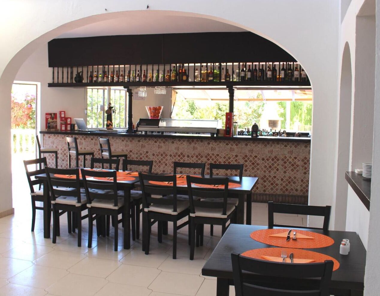 Ein Restaurant mit Fliesenboden, Holztischen und -stühlen, einer Bar mit Flaschen und Blick durch große Fenster.