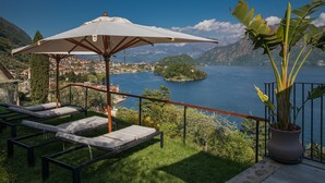 Eine Terrasse mit Liegestühlen und einem großen Sonnenschirm mit Blick auf einen See und Berge.