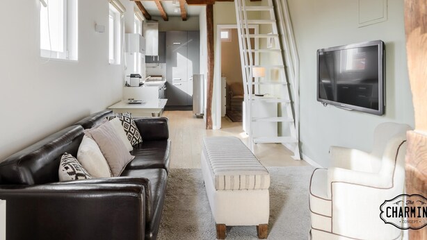 Un salon moderne avec un canapé en cuir noir, un pouf blanc et une chaise blanche. Un téléviseur est fixé au mur, et un escalier en bois mène à un niveau supérieur.
