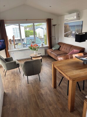 Una sala moderna con un sofá de cuero marrón, una mesa de centro de madera y una mesa de comedor. Hay un ventanal con vistas al exterior, un radiador y un aire acondicionado instalado.