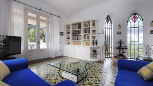 Ein Wohnzimmer mit einem blauen Sofa, einem Glastisch und einem Fliesenboden. Bücherregale sind mit Büchern und Dekorationsgegenständen gefüllt.