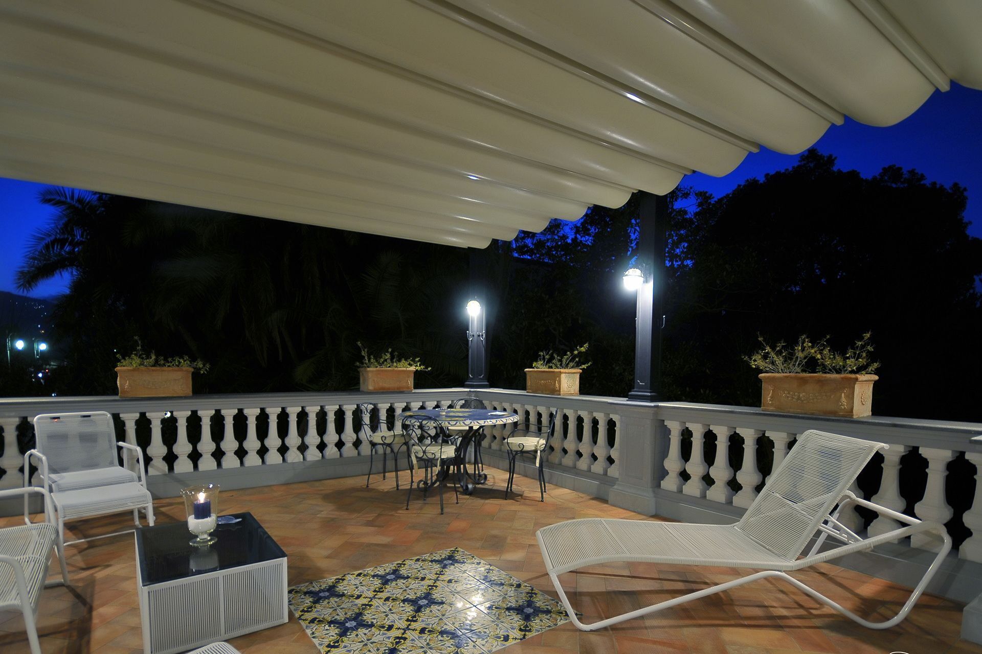 Un patio al aire libre con una zona de estar cubierta, que incluye sillas y una mesa, rodeado de plantas en macetas y un piso con diseño.