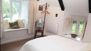 A bedroom with a large bed, a window seat with cushions, a chair, and a coat rack. - A beautiful 250 year old thatched cottage close to Frenchman's Creek. 2 bedrooms (Helston)