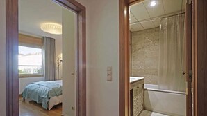 A bedroom with a bed, a window with curtains, and a bathroom with a shower and a marble floor. - GREEN ÁVILA (LISBOA)