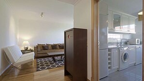 A compact living space with a sofa, a small table, a dark-colored cabinet, and a kitchen area with a washing machine. - GREEN ÁVILA (LISBOA)