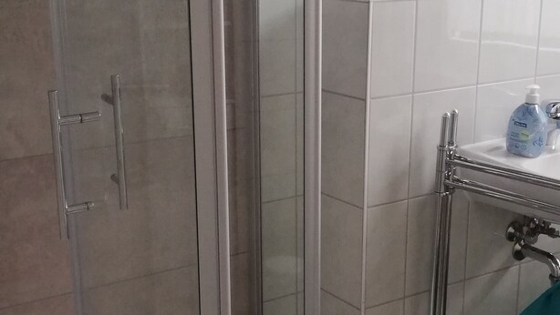 Un espace douche doté d’une porte en verre, d’une barre d’appui et d’une petite étagère sur laquelle se trouve une bouteille.