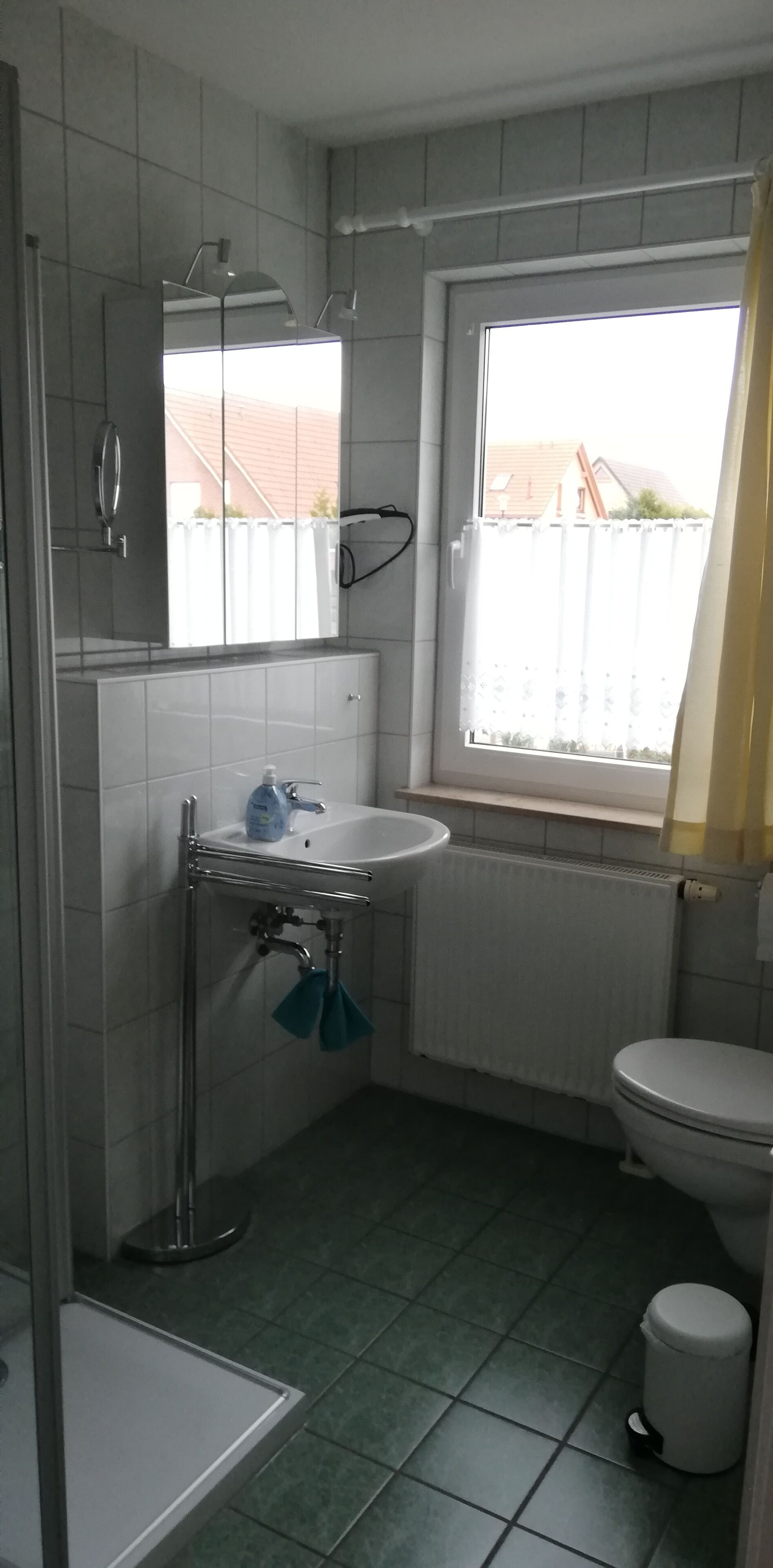 Ein kleines Badezimmer mit einem weißen Waschbecken, einem Spiegel darüber, einer Toilette und einem Fenster mit Blick auf einen Zaun und Gebäude außerhalb.