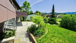 Ein Wohngebäude mit überdachter Außenfläche, einer Treppe und Blick auf eine grüne Landschaft mit Bäumen und Hügeln.