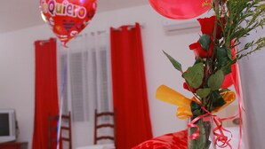 Ein Zimmer mit einem Bett, einem Nachttisch mit einer Schale Nüsse, einer Vase mit Rosen und roten Ballons, darunter ein Herzballon mit der Aufschrift 'Te Quiero'.