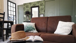 A brown sofa with a palm leaf pillow and a white pillow with text, a wicker hat, and an open book on a small table. - 2-bedroom house, close to the beach, all on foot (L'aiguillon-Sur-Mer)