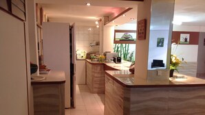 Eine moderne Küche mit Marmorarbeitsplatte, einem eingebauten Ofen und einem Spülbecken. Links befinden sich ein kleiner Mülleimer und ein Kühlschrank. Die Küche verfügt über eine geflieste Spritzschutzwand und ein Fenster mit Blick nach draußen.