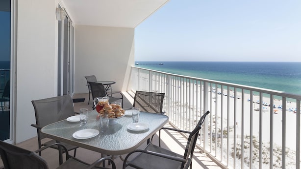 A balcony with a table set for dining, overlooking a beach with a pier in the distance. - Beach Front Condo, Spacious, Clean and Comfortable (Gulf Shores)