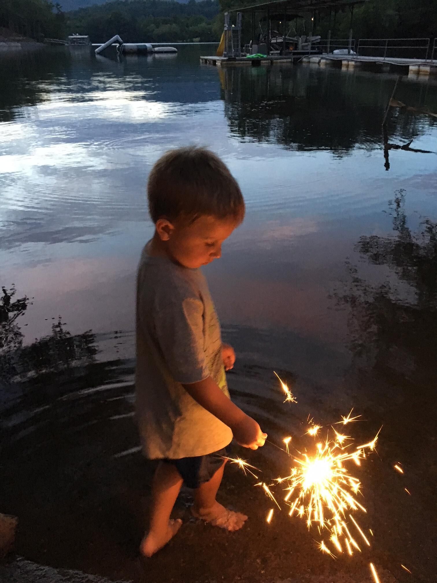 Un niño sosteniendo una bengala cerca del agua, con un muelle y estructuras industriales al fondo.