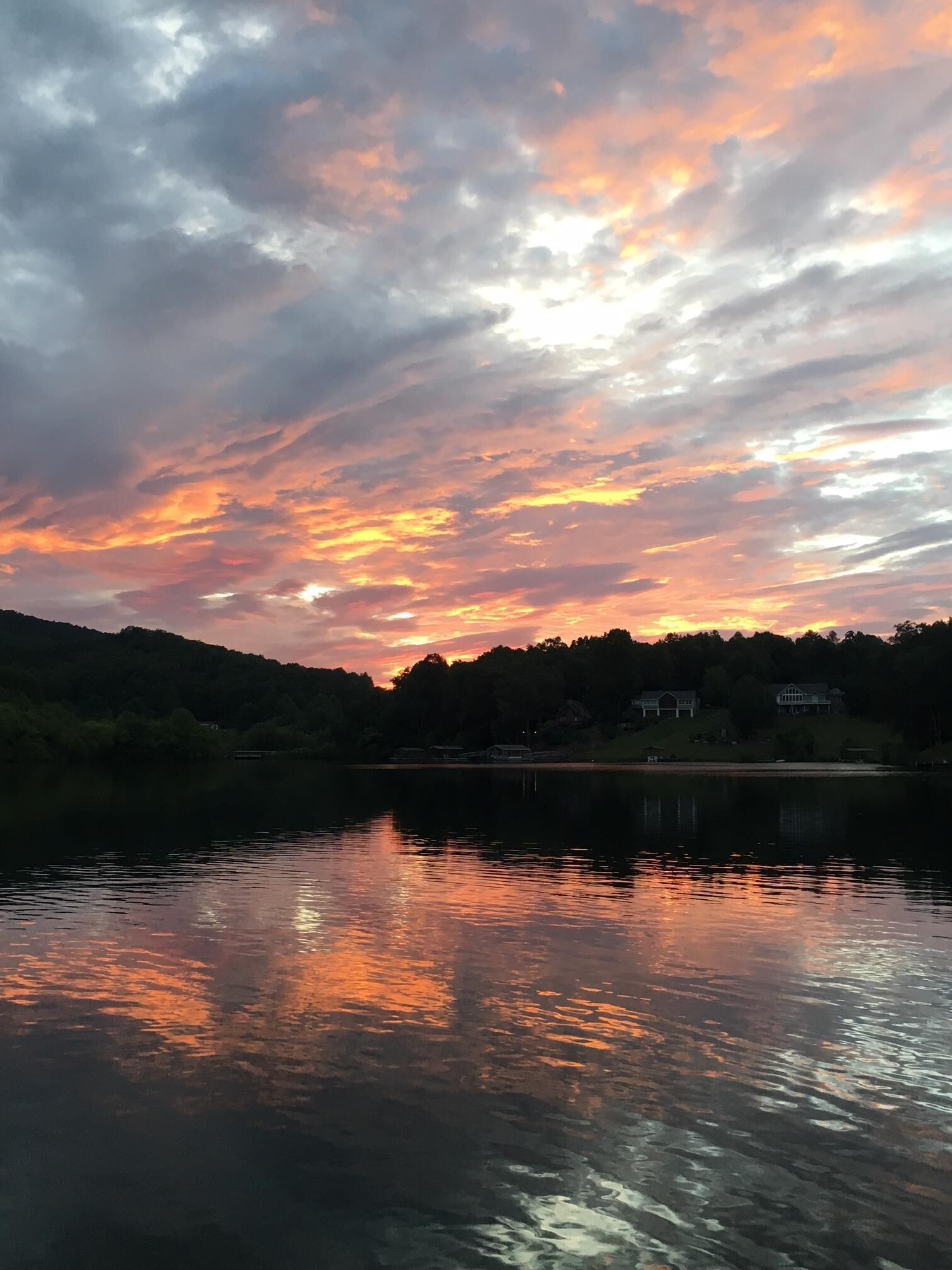 A calm lake with a sunset reflection, surrounded by a forested area and houses in the background.