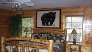 A log cabin bedroom with a wooden bed, a framed bear picture, two lamps, and a window with blinds. - Big Nate Cabin on Lake with Water Trampoline (Hiawassee)
